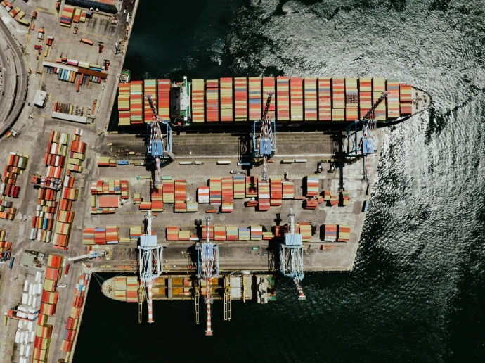 Top-down aerial view of a busy container port terminal with cargo ships docked and cranes loading colorful shipping containers.