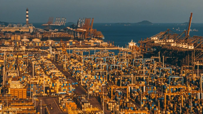 Wide-angle view of a massive container terminal at sunset, showing stacked cargo containers and port cranes along the coastline.