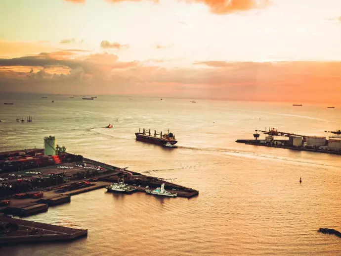High-angle view of cargo ships navigating a busy port channel at sunset, featuring coastal industrial terminals and various vessels on the open water.